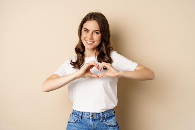 Belle femme montrant coeur, signe d'amour et souriant, debout sur fond beige en t-shirt blanc, fond beige.