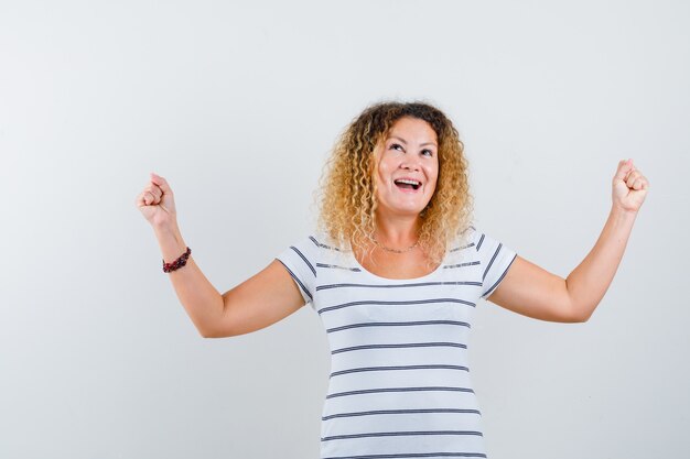 Belle femme montrant le geste du gagnant en t-shirt et ayant l'air réussie. vue de face.