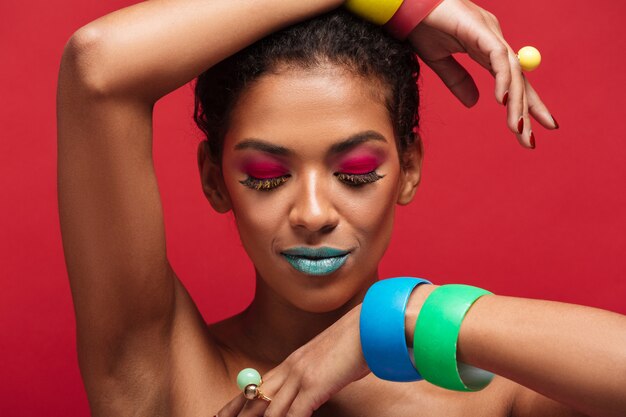 Belle femme mulâtre à moitié nue avec un maquillage tendance démontrant des bracelets colorés sur l'appareil photo, sur le mur rouge