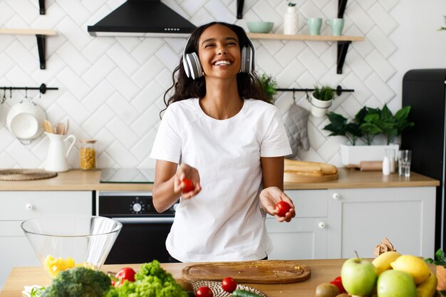 Belle femme mulâtre sourit tient des tomates et écoute quelque chose dans de gros écouteurs près de la table pleine de légumes frais dans la cuisine moderne vêtue d'un t-shirt blanc