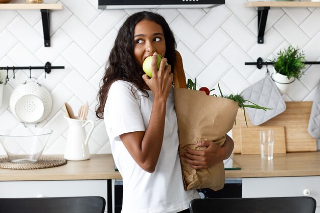 Belle femme mulâtre tient le paquet plein de nourriture et mange des pommes sur la cuisine blanche moderne