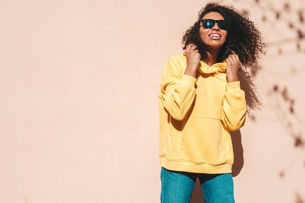 Belle femme noire avec des boucles afro coiffureModèle hipster souriant en sweat à capuche jaune Sexy femme insouciante posant sur le fond de la rue en lunettes de soleil Enthousiaste et heureux