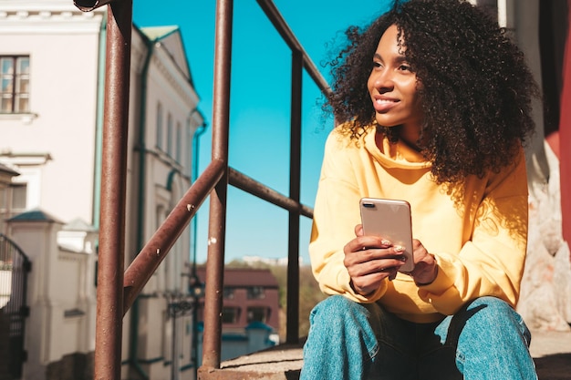 Belle femme noire avec des boucles afro coiffureModèle souriant en sweat à capuche jaune Sexy femme insouciante posant sur le fond de la rue à lunettes de soleil en regardant l'écran du smartphone à l'aide d'applications