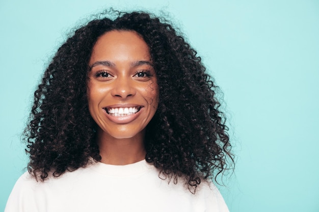 Photo gratuite belle femme noire avec une coiffure afro curls modèle souriant en vêtements longs t-shirt sexy femme insouciante posant près du mur bleu en studio bronzé et joyeux