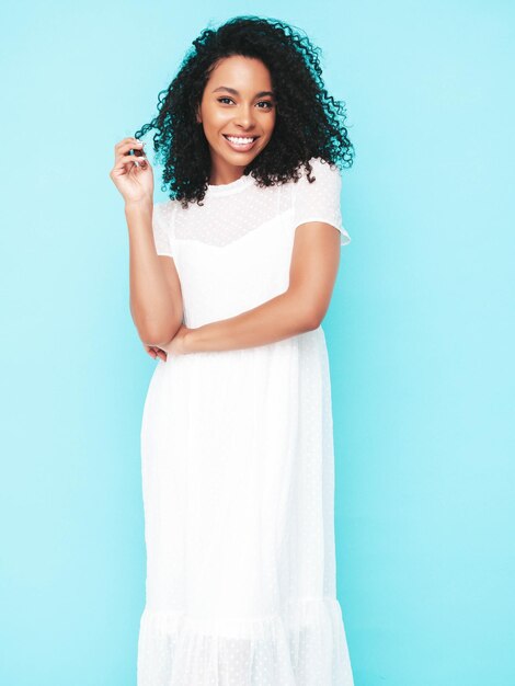 Belle femme noire avec une coiffure afro curls Modèle souriant vêtu d'une robe d'été blanche Sexy femme insouciante posant près du mur bleu en studio Bronzé et joyeux