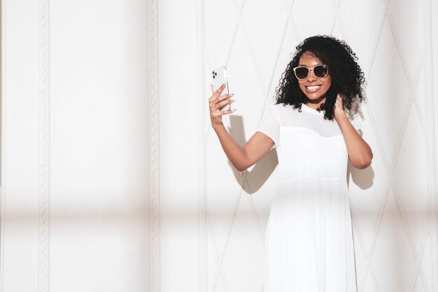 Belle femme noire avec une coiffure afro curls Modèle souriant vêtu d'une robe d'été blanche Sexy femme insouciante posant près du mur en studioAu jour ensoleillé Ombre de la fenêtre Prendre selfie dans les lunettes