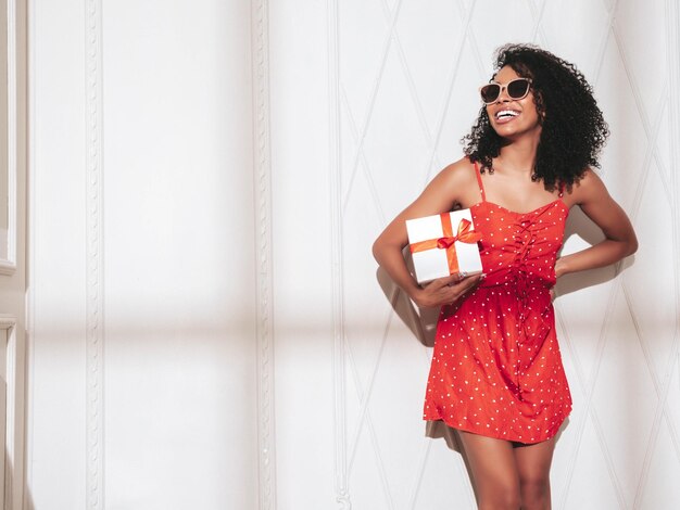 Belle femme noire avec une coiffure afro curls Modèle souriant vêtu d'une robe d'été rouge Sexy femme insouciante posant près d'un mur blanc en studio Bronzé et joyeux tenant une boîte-cadeau