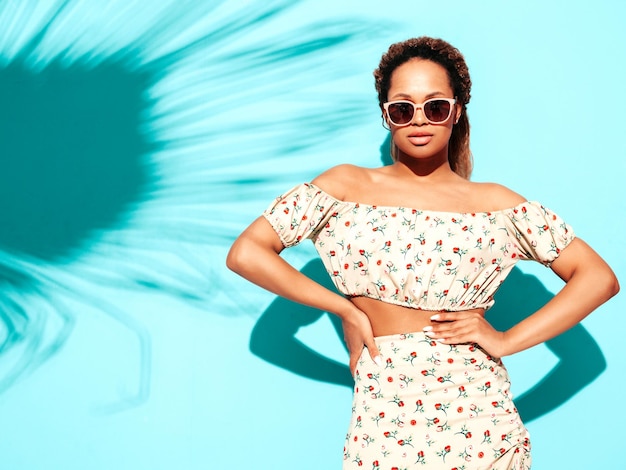 Belle femme noire avec une coiffure afro curls Modèle souriant vêtu de vêtements hipster d'été Sexy femme insouciante posant près du mur bleu en studio Bronzé et joyeux