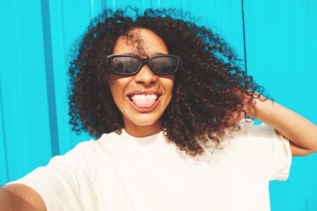 Belle femme noire avec une coiffure afro curlsModèle hipster souriant en t-shirt blancFemme insouciante sexy posant dans la rue près du mur bleu en lunettes de soleilGai et heureuxPrendre une photo de selfie Pov