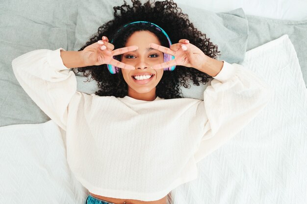 Belle femme noire avec une coiffure de boucles afro. Modèle souriant en pull et jeans. femme insouciante écoutant de la musique dans des écouteurs sans fil le matin