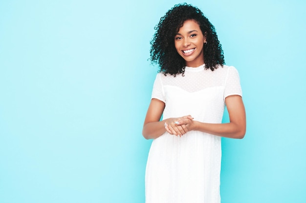 Belle femme noire avec une coiffure de boucles afro Modèle souriant vêtu d'une robe d'été blanche Sexy femme insouciante posant près du mur bleu en studio Bronzé et joyeux Isolé