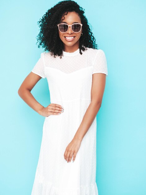 Belle femme noire avec une coiffure de boucles afro Modèle souriant vêtu d'une robe d'été blanche Sexy femme insouciante posant près du mur bleu en studio Bronzé et joyeux En lunettes de soleil Isolé