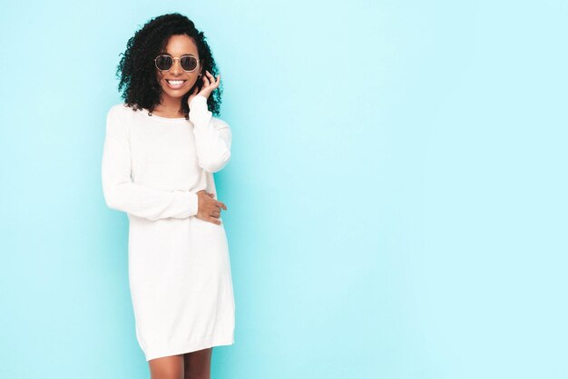 Belle femme noire avec une coiffure de boucles afro Modèle souriant vêtu d'une robe d'été blanche Sexy femme insouciante posant près du mur bleu en studio Bronzé et joyeux En lunettes de soleil Isolé