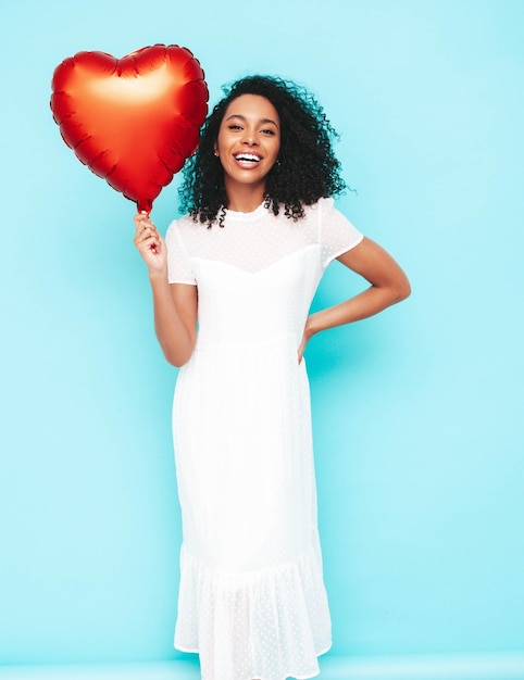 Belle femme noire avec une coiffure de boucles afro Modèle souriant vêtu d'une robe d'été blanche Sexy femme insouciante posant près du mur bleu en studio Bronzé et joyeux Tenant un ballon à air coeur