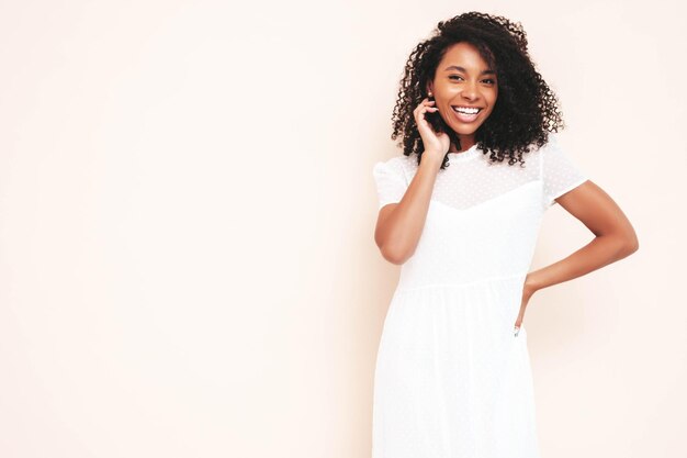 Belle femme noire avec une coiffure de boucles afro Modèle souriant vêtu d'une robe d'été blanche Sexy femme insouciante posant près du mur en studio Bronzé et joyeux Isolé