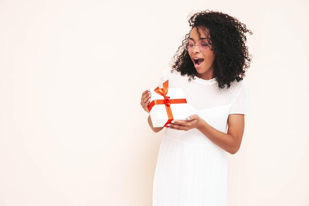 Belle femme noire avec une coiffure de boucles afro Modèle souriant vêtu d'une robe d'été blanche Sexy femme insouciante posant près du mur en studio Bronzé et joyeux Tenant une boîte-cadeau Isolé