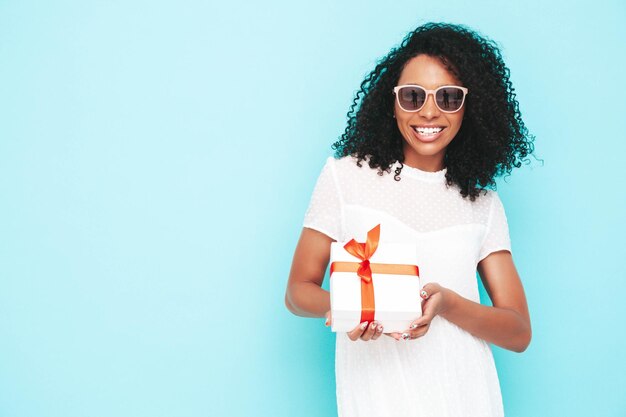 Belle femme noire avec une coiffure de boucles afro Modèle souriant vêtu d'une robe d'été blanche Sexy femme insouciante posant près d'un mur bleu en studio Bronzé et joyeux Tenant une boîte-cadeau Isolé