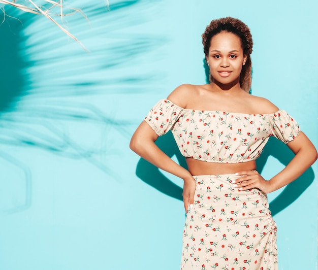 Belle femme noire avec une coiffure de boucles afro Modèle souriant vêtu de vêtements hipster d'été Sexy femme insouciante posant près du mur bleu en studio Bronzé et joyeux