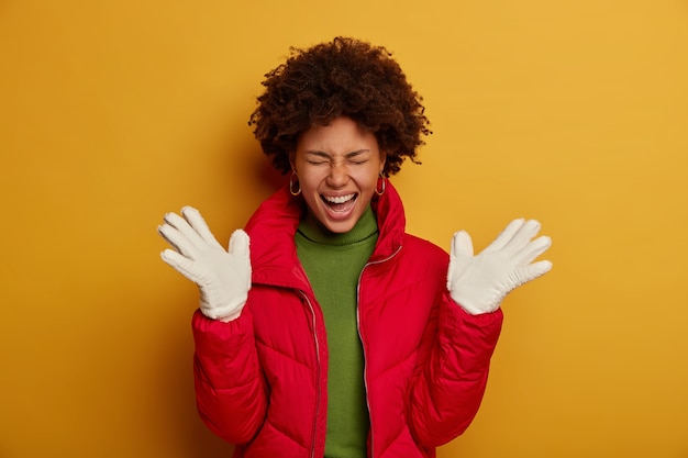 Photo gratuite belle femme à la peau foncée aux cheveux bouclés, porte des vêtements d'extérieur d'hiver, des gants blancs, exprime le bonheur, s'exclame de plaisir, isolée sur le mur jaune du studio.