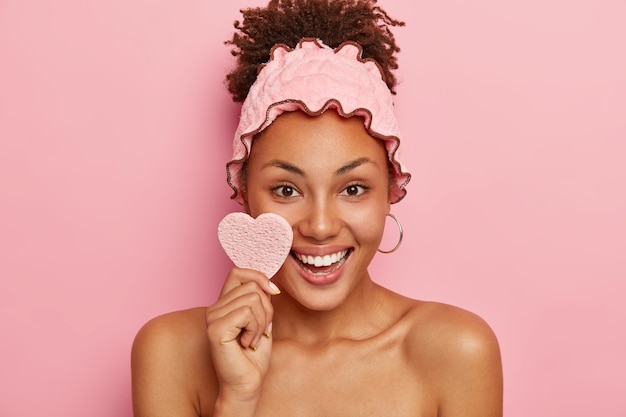 Belle femme a une peau saine, essuie le visage avec une éponge cosmétique, sourit positivement, montre des dents blanches, porte un bandeau de douche, prend une douche après le travail, isolé sur un mur rose