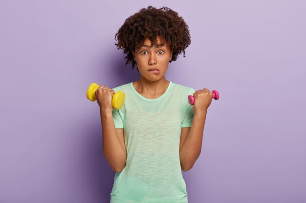 Belle femme à la peau sombre mord les lèvres avec un regard minutieux, soulève deux haltères, balance les biceps