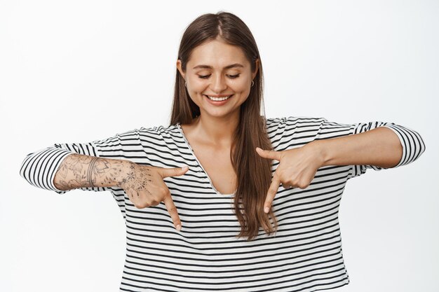 Belle femme pointant et regardant vers le bas avec un sourire heureux, debout dans un chemisier rayé décontracté sur fond blanc.