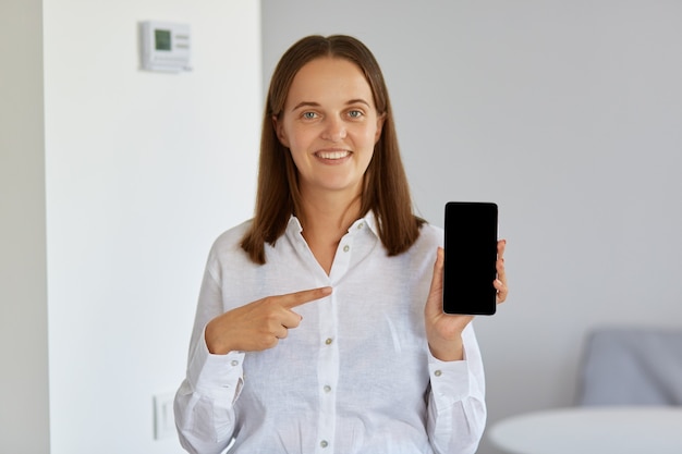 Belle femme portant une chemise blanche tenant un téléphone intelligent dans les mains et pointant vers son écran blanc avec l'index, regardant la caméra avec un sourire charmant.