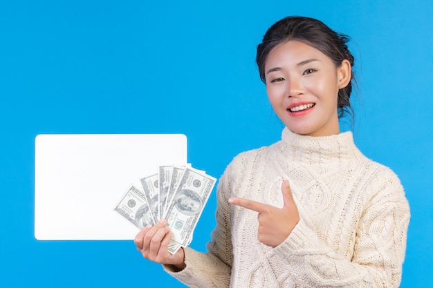 Une belle femme portant un nouveau tapis blanc à manches longues tenant une pancarte blanche et un billet d'un dollar bleu. Commerce .