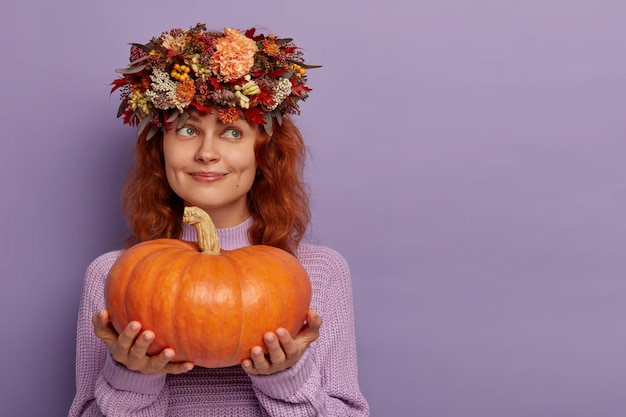 Belle femme rêveuse tient une grosse citrouille mûre, regarde pensivement loin, vêtue d'un pull surdimensionné, des modèles de guirlande contre un mur violet