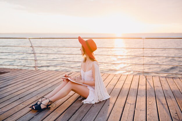 Belle femme en robe blanche assise au bord de la mer au lever du soleil en pensant et en prenant des notes