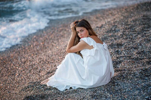 Belle femme en robe blanche assise seule et regardant en bord de mer.