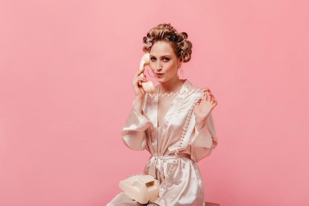 Belle femme en robe de chambre en soie parlant avec coquetterie au téléphone sur un mur rose
