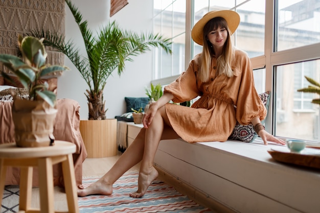 Belle femme en robe de lin et chapeau de paille posant dans un appartement de style bohème