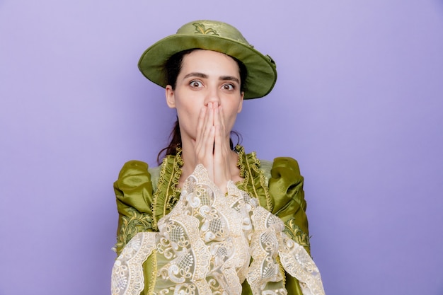 Photo gratuite belle femme en robe renaissance et chapeau choqué couvrant la bouche avec les mains sur le bleu