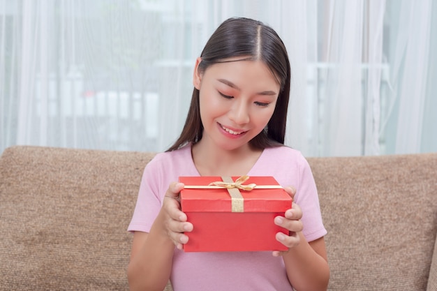 Belle femme en robe rose, assise sur le canapé, ouvrant une boîte-cadeau.