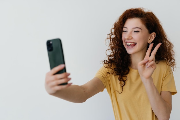 Belle femme rousse aux cheveux bouclés touchant sa coiffure souriante et prenant selfie faisant photo sur smartphone sur fond blanc