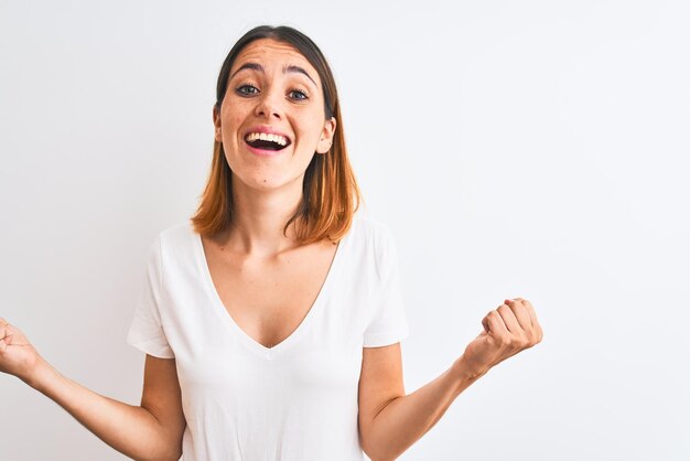 Belle femme rousse portant un t-shirt blanc décontracté sur fond isolé célébrant la surprise et l'émerveillement du succès avec les bras levés et les yeux ouverts Concept gagnant