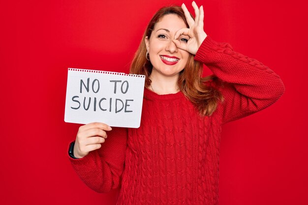 Belle femme rousse tenant une bannière montrant non au message de suicide sur fond rouge avec un visage heureux souriant faisant signe ok avec la main sur les yeux regardant à travers les doigts