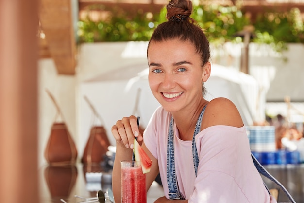 Belle femme satisfaite avec un sourire agréable, boit un cocktail, s'assoit à la cafétéria, a un chignon et un sourire éclatant, étant heureuse de passer des vacances dans un pays tropical.