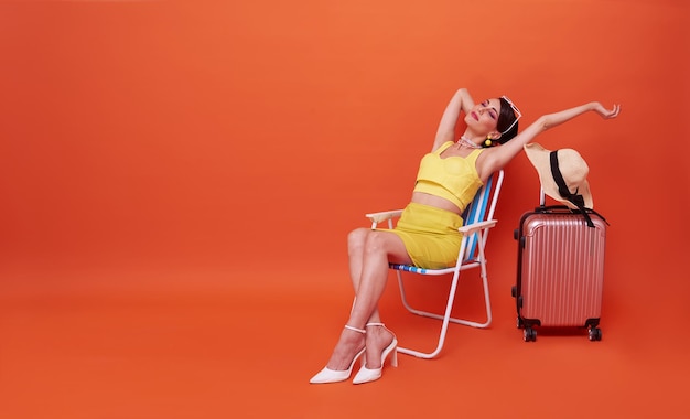 Photo gratuite belle femme se détendre assis sur une chaise de plage avec une valise en studio été fond orange