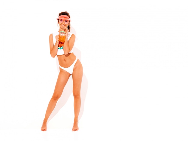 Belle femme sexy souriante en slip d'été blanc et sujet. Fille branchée en lunettes de soleil et casquette de visière transparente Devenir fou. Modèle drôle isolé. Boire une boisson fraîche de cocktail