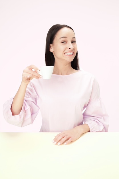 Belle Femme Souriante Assise Au Studio Rose Et Regardant Heureux Tenant La Tasse De Café à La Main. Closeup Portrait Tonique Dans Un Style Minimalisme