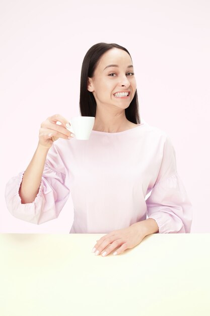 Belle femme souriante assise au studio rose et regardant heureux tenant la tasse de café à la main. Closeup portrait tonique dans un style minimalisme