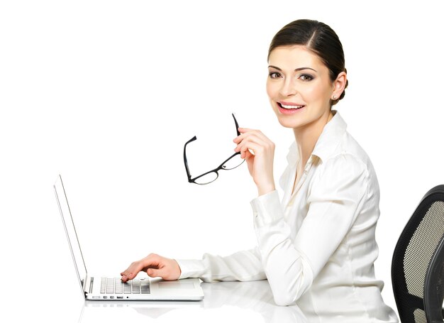 Belle femme souriante est assise de la table et travaille sur un ordinateur portable en chemise blanche - isolé sur blanc.