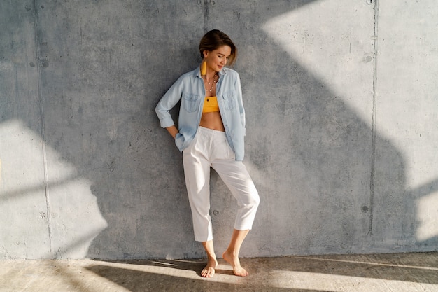 Belle Femme Souriante Et Heureuse Posant Contre Un Mur De Béton Par Une Journée Ensoleillée Portant Une Chemise En Jean, Un Haut Jaune, Un Pantalon Blanc