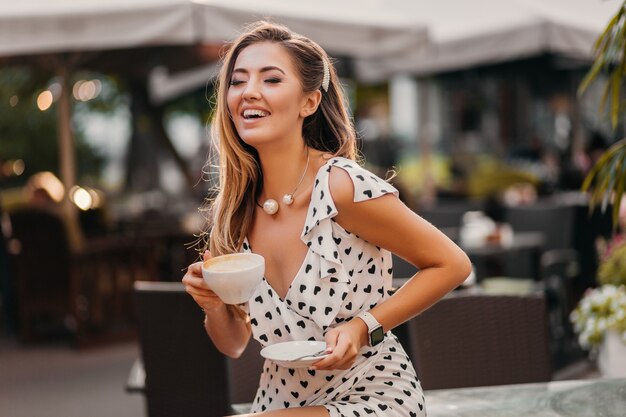 Belle femme souriante portant une robe imprimée blanche élégante assis dans un café de rue avec une tasse de cappuccino