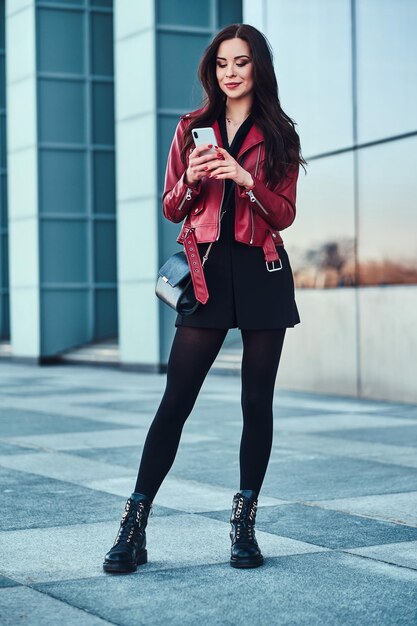 Belle femme souriante en veste rouge se tient à côté d'un bâtiment en verre et discute avec quelqu'un par téléphone portable.