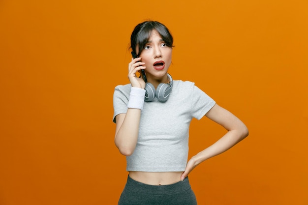 Belle femme sportive en tenue de sport avec un casque parlant sur un téléphone portable à la confusion debout sur fond orange