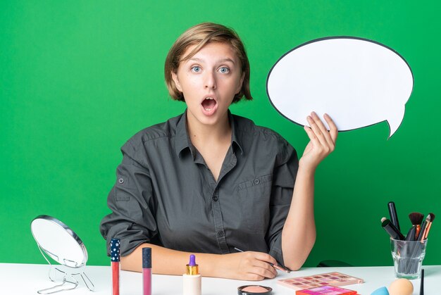 Une belle femme surprise est assise à table avec des outils de maquillage tenant une bulle de dialogue avec un pinceau de maquillage