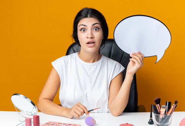 Une belle femme surprise est assise à table avec des outils de maquillage tenant une bulle de dialogue avec un pinceau de maquillage